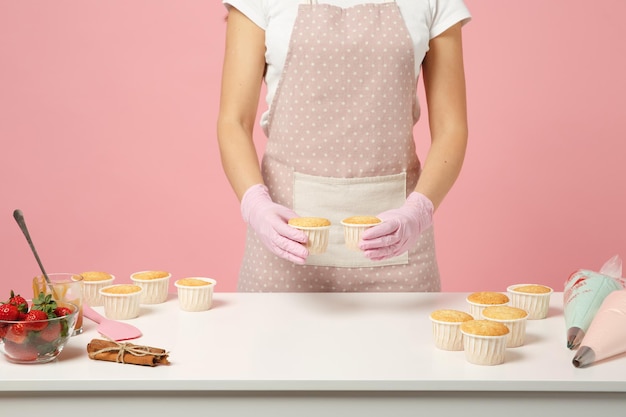 Feche o cozinheiro chefe de dona de casa bonito recortado confeiteiro ou padeiro em camiseta branca cozinhando na mesa isolada em fundo rosa pastel no estúdio. processo de fabricação de cupcakes. mock up conceito de comida de espaço de cópia.