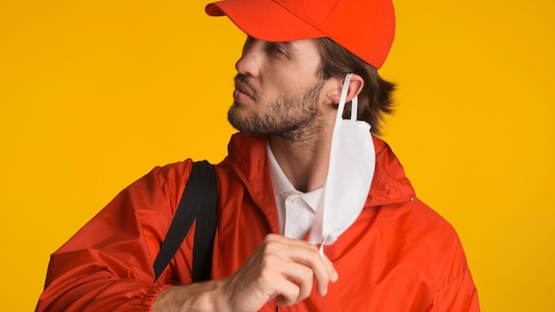 Feche o entregador vestido de uniforme tirando máscara facial parecendo cansado depois do dia de trabalho Correio masculino em pé sobre fundo colorido