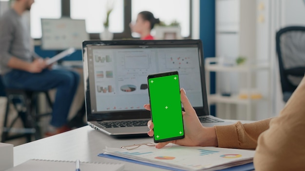 Feche o funcionário segurando verticalmente a tela verde no smartphone no escritório de negócios. Mulher que trabalha com modelo de simulação isolado e fundo de croma no visor do celular.