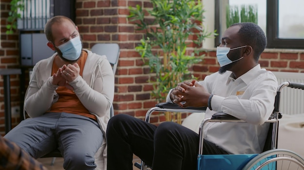 Feche o homem afro-americano sentado em cadeira de rodas em uma reunião de grupo, usando máscara facial. Adulto com deficiência participando de sessão de terapia com pessoas durante a pandemia de covid 19.