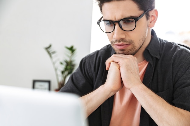 Feche o homem sério e bonito em óculos usando laptop enquanto está sentado à mesa no escritório