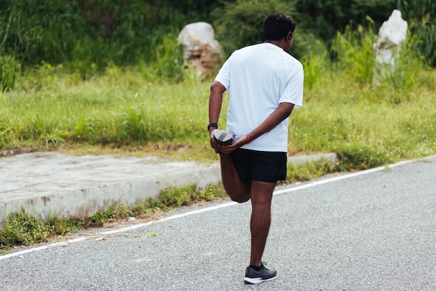 Feche o jovem atleta asiático, corredor esportivo, homem negro, use relógio, levante os pés, esticando as pernas e o joelho antes de correr no parque de saúde de rua ao ar livre, exercício saudável antes do conceito de treino