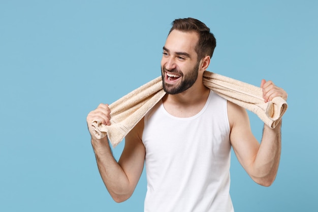 Feche o jovem barbudo de 20 anos de idade na camisa branca, segure a toalha nos ombros, isolada no retrato de estúdio de fundo pastel azul. Conceito de procedimentos cosméticos de saúde de cuidados com a pele. Simule o espaço da cópia