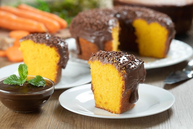 Feche o pedaço de bolo de cenoura brasileiro com cobertura de chocolate na mesa de madeira com cenouras ao fundo