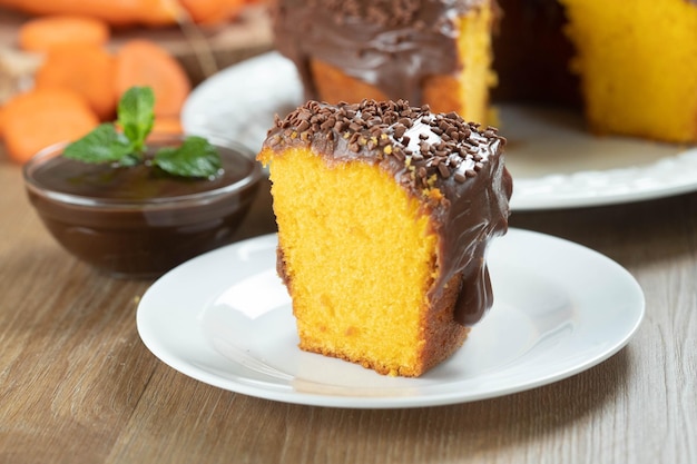 Feche o pedaço de bolo de cenoura brasileiro com cobertura de chocolate na mesa de madeira com cenouras ao fundo