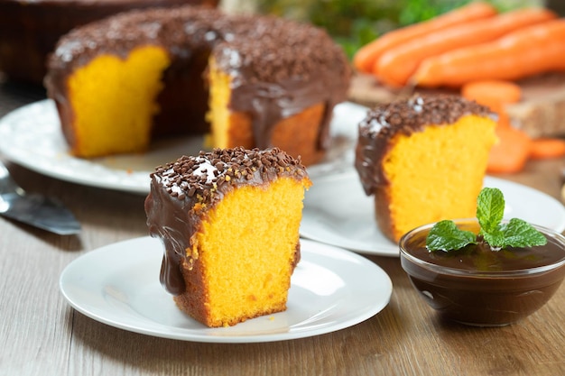 Feche o pedaço de bolo de cenoura brasileiro com cobertura de chocolate na mesa de madeira com cenouras ao fundo