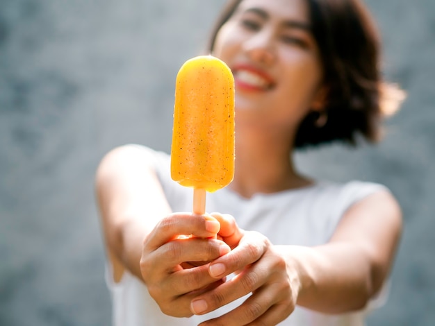 Feche o picolé amarelo na mão de linda mulher asiática feliz vestindo camisa branca sem mangas casual, ao ar livre. Mulher mostrando picolés. Mulher sorridente, desfrutando de picolé no verão.