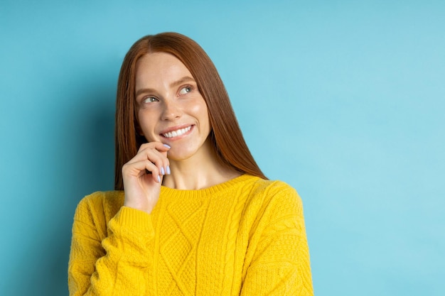 Feche o retrato da adorável feliz misteriosa ruiva frecled modelo feminino sem maquiagem, pensando em algo agradável, olhando de lado sorrindo contra o fundo azul do estúdio.