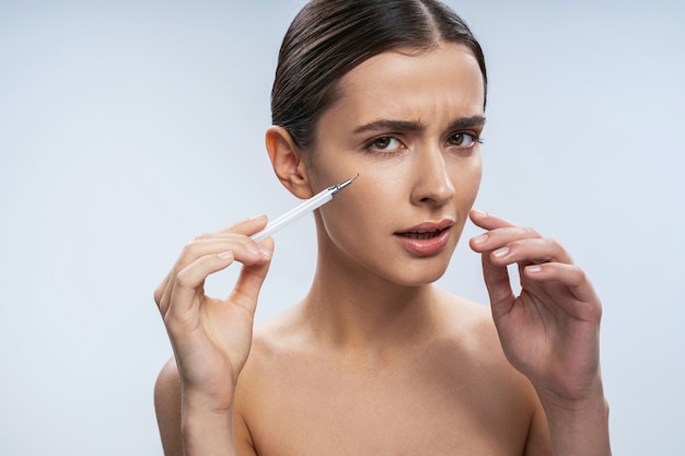 Feche o retrato da elegante mulher branca fazendo uma injeção de ácido hialurônico sob os olhos, isolado no fundo branco