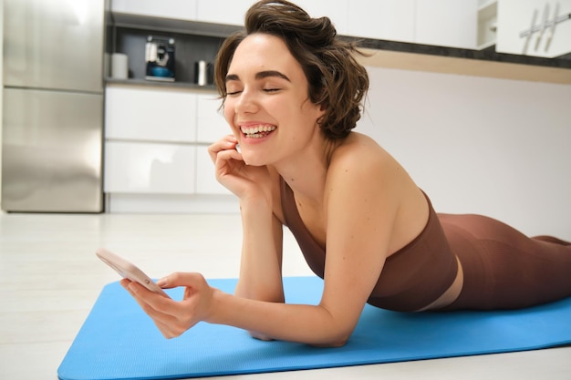 Feche o retrato da garota fitness deitada no tapete de ioga e rindo usando o ex de treino de registros de smartphone