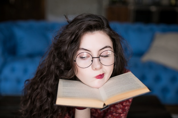 Feche o retrato da menina atraente estudante com cabelos cacheados em óculos redondos. livro de leitura jovem nas mãos perto do rosto, coloque o queixo nele. suas sobrancelhas e lábios expressam ceticismo e descrença