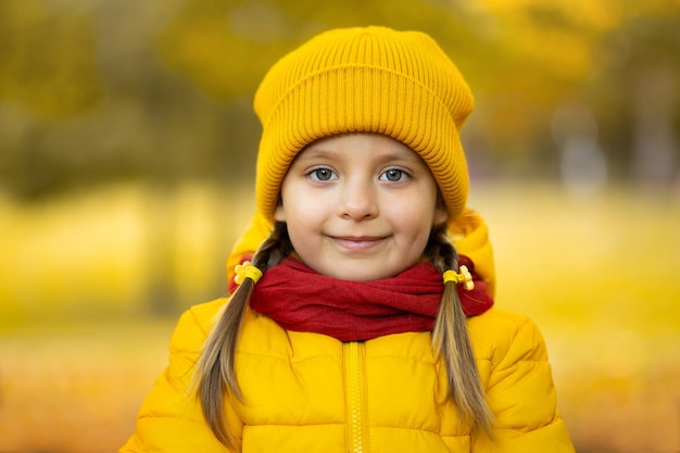 Feche o retrato da menina bonita bonito na tampa amarela, jaqueta e lenço vermelho, sorrindo para a câmera em fundo dourado lindo de outono ao ar livre no parque.