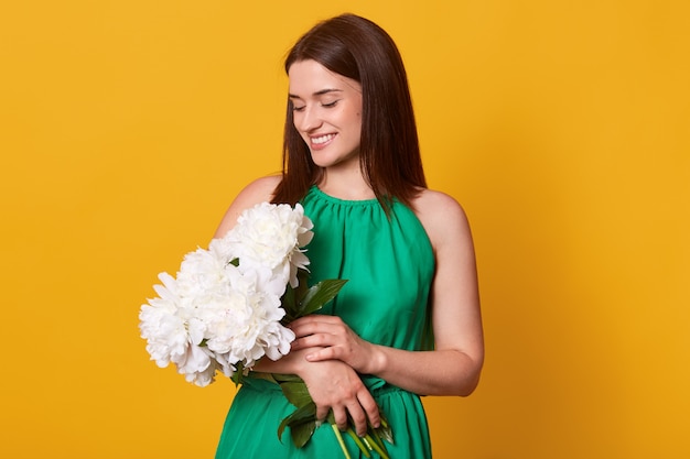 Feche o retrato da mulher elegante atraente na data em elegante vestido verde na parede amarela segurando o buquê de flores de peônia, tem expressão facial agradável, cai feliz. Conceito de amor