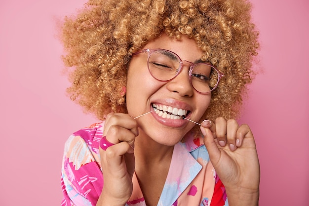 Feche o retrato da mulher europeia de cabelos cacheados limpa os dentes com fio dental pisca o olho usa óculos transparentes e camisa colorida isolada sobre fundo rosa conceito de higiene oral