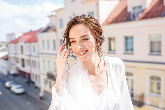 Feche o retrato da noiva linda e feliz em um peignoir ficar em uma varanda e espera o noivo no dia do casamento