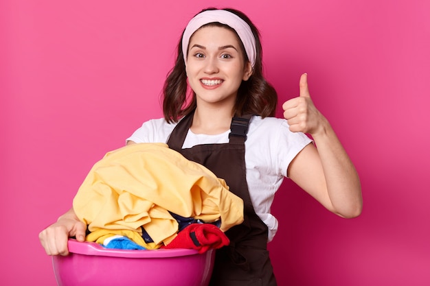 Feche o retrato de dona de casa esbelta bonita segurando a bacia rosa com roupas limpas, com expressão facial positiva, mostrando super sinal com o punho e o polegar, parece emocional e feliz.