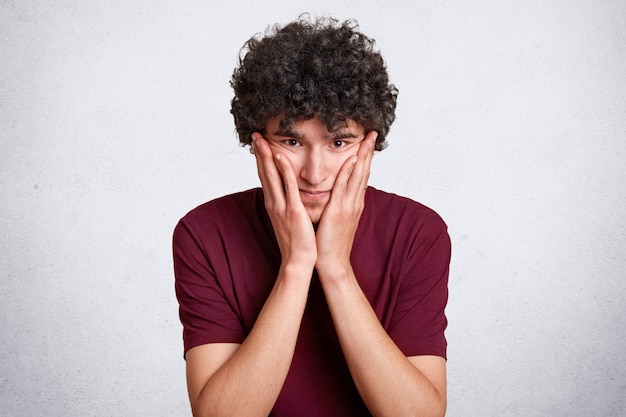 Feche o retrato de homem sério toca a bochecha com a palma da mão, olha diretamente para a câmera, tem expressão pensativa, veste camiseta marrom casual