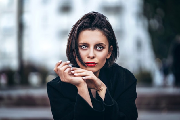 Feche o retrato de jovem com corte de cabelo curto e lábios vermelhos, vestido de preto. Mulher posando em uma cidade urbana turva