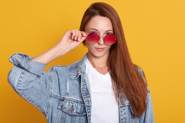 Feche o retrato de jovem na camisa branca usa óculos escuros e jaqueta jeans. garota atraente com cabelo escuro posa com as mãos