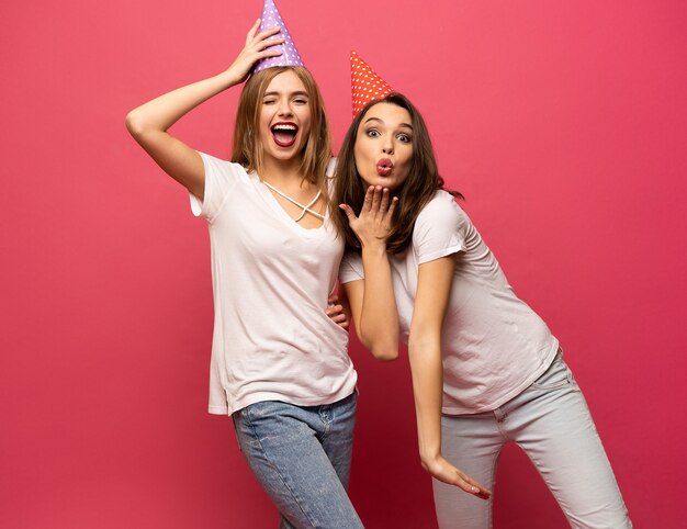 Feche o retrato de mulheres jovens loiras e morenas com chapéus de aniversário se divertindo isolado no fundo rosa.