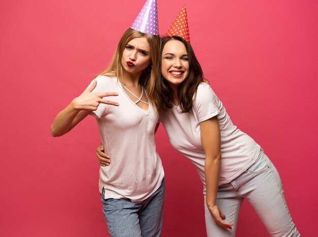 Feche o retrato de mulheres jovens loiras e morenas com chapéus de aniversário se divertindo isolado no fundo rosa.