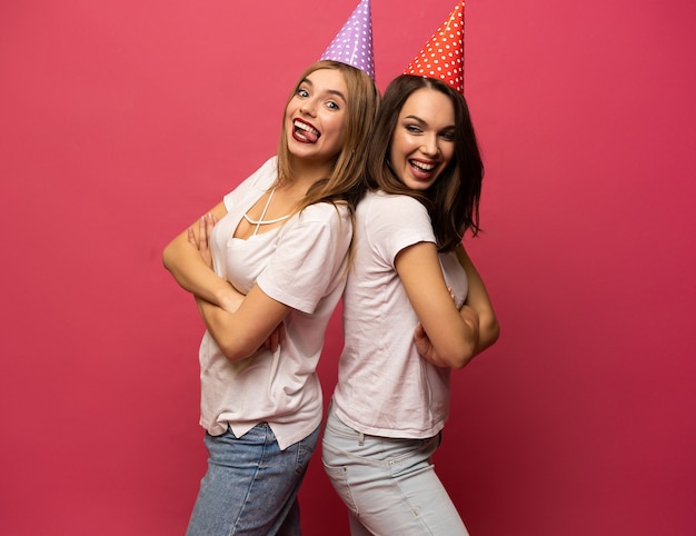 Feche o retrato de mulheres jovens loiras e morenas com chapéus de aniversário se divertindo isolado no fundo rosa.