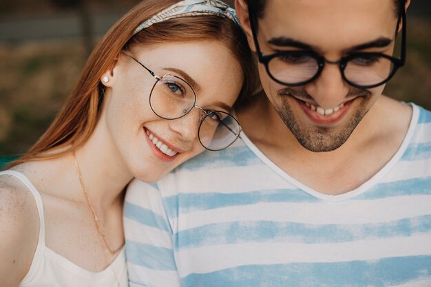 Foto feche o retrato de um adorável jovem casal namorando enquanto a mulher está olhando para a câmera sorrindo, inclinando a cabeça no namorado, que está sorrindo ao ar livre.