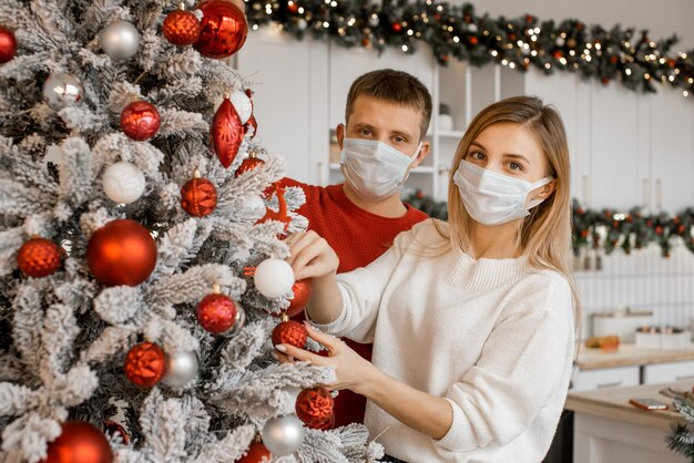 Feche o retrato de um casal de família usando máscara médica protetora de rosto para proteção contra doenças virais, decorando a árvore de Natal em casa e olhando para a câmera