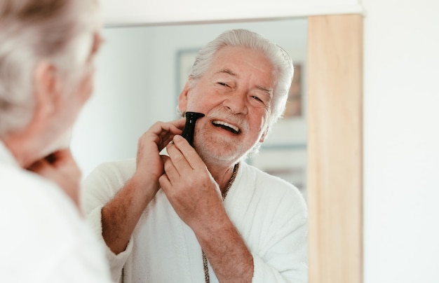 Feche o retrato de um homem bonito sênior em roupão de barbear barba com um barbeador elétrico olhando para si mesmo na frente do espelho