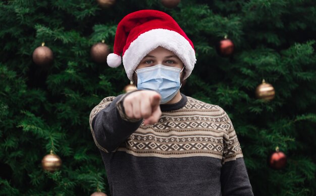 Feche o retrato de um homem com um chapéu de Papai Noel e uma máscara médica com emoção e mostre o dedo. No contexto de uma árvore de Natal. Pandemia do coronavírus