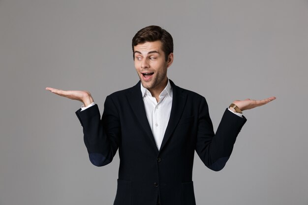 Feche o retrato de um jovem empresário alegre vestido com um terno isolado sobre uma parede cinza, apresentando espaço de cópia nas palmas das mãos.