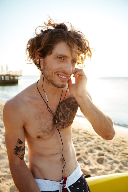 Feche o retrato de um jovem surfista cacheado bonito ouvindo música com fones de ouvido enquanto está sentado na prancha de surf na praia