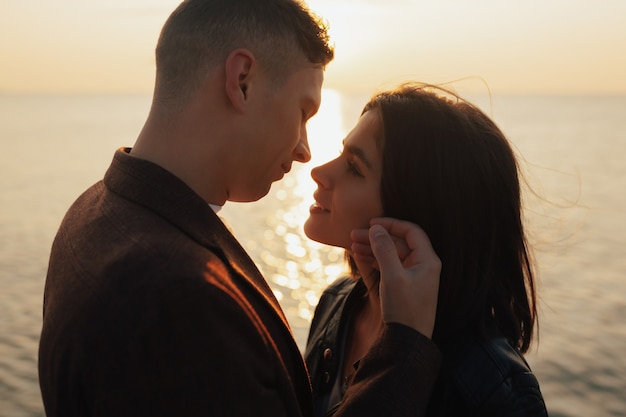 Feche o retrato de um lindo casal caucasiano olhando um ao outro e sorrindo enquanto o homem está tocando o rosto da namorada ao pôr do sol do mar.