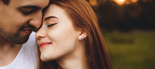 Feche o retrato de um lindo casal jovem sentado cara a cara com os olhos fechados, sorrindo antes de beijar, enquanto o homem a abraça de costas contra o pôr do sol.