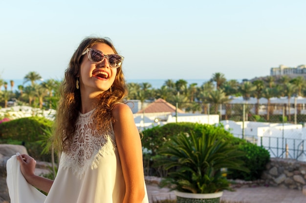 Feche o retrato de uma adolescente de vestido branco e óculos de sol em um parque durante um dia ensolarado olhando e sorrindo contra o céu e o mar