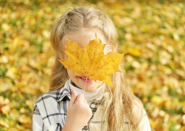 Feche o retrato de uma adorável criança lúdica se divertindo em uma caminhada no belo parque de outono posando na câmera com folhas amarelas cobrindo seu rosto Foto horizontal