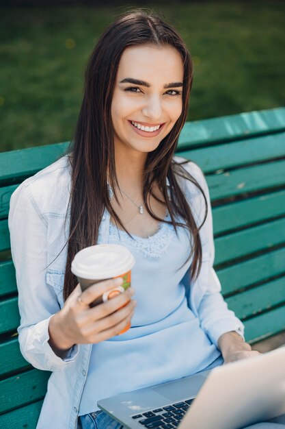 Feche o retrato de uma bela jovem com cabelo escuro, sorrindo, segurando uma xícara de café e um laptop com as pernas, sentado em um banco do lado de fora.