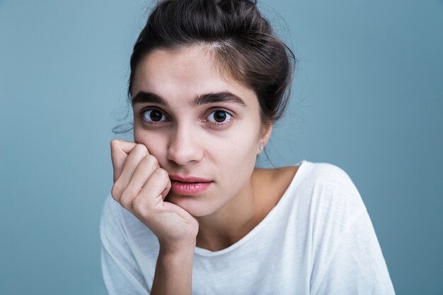 Feche o retrato de uma bela jovem morena vestindo uma camiseta branca em pé, isolado sobre um fundo azul, posando, tocando o rosto