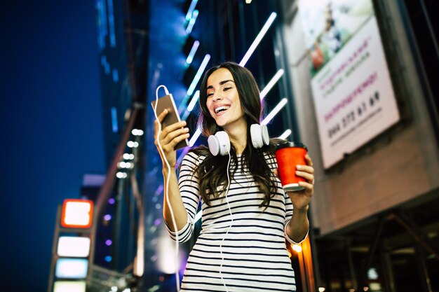 Feche o retrato de uma jovem atraente e feliz em fones de ouvido com telefone inteligente nas mãos ao ar livre