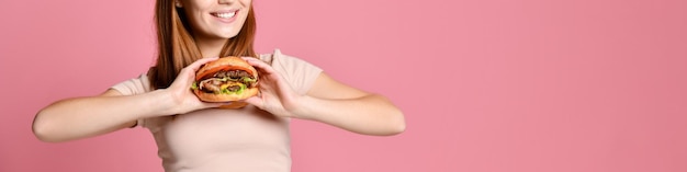 Feche o retrato de uma jovem faminta comendo hambúrguer isolado sobre fundo rosa