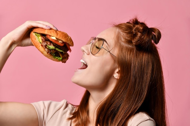 Feche o retrato de uma jovem faminta comendo hambúrguer isolado sobre fundo rosa