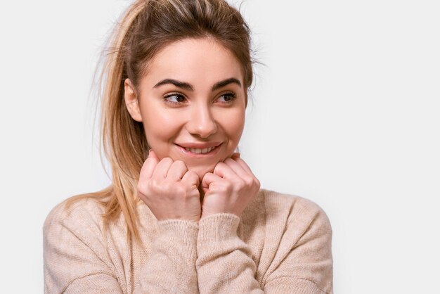 Foto feche o retrato de uma jovem gentil e fofa sorrindo e mantém as mãos no queixo, olhando de lado a camisola bege vestida isolada sobre o fundo branco pessoas emoções sentimento e conceito de linguagem corporal