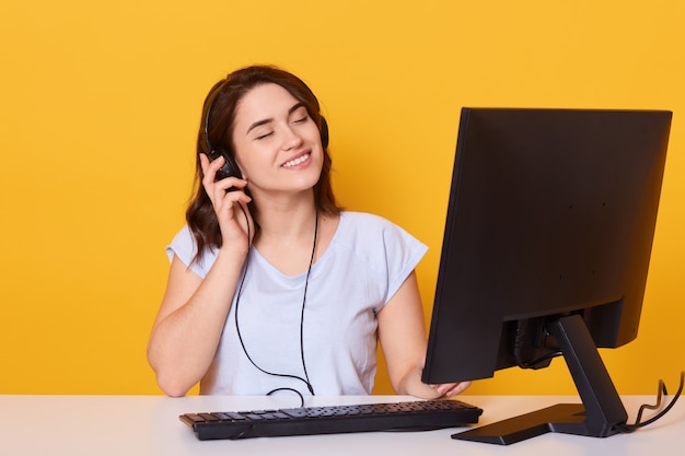 Feche o retrato de uma jovem mulher atraente, sentado à mesa com o computador e ouvir música, posando isolado sobre amarelo