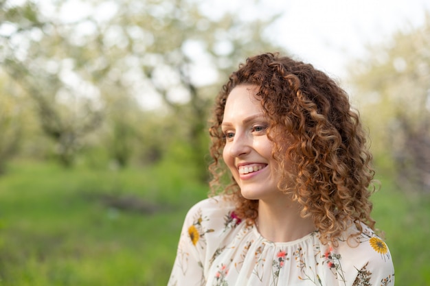 Feche o retrato de uma jovem mulher atraente sorridente com cabelos cacheados no parque primavera floração verde. Emoções puras.