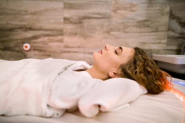 Feche o retrato de uma jovem mulher bonita, deitado na cama especial spa quente, coberto com manta quente durante o procedimento de quebra de lama no spa.