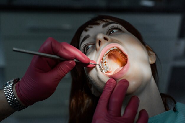 Feche o retrato de uma jovem paciente do sexo feminino com a boca aberta, com tratamento odontológico profissional e as mãos do médico usando luvas de borracha vermelha, segurando o espelho dental