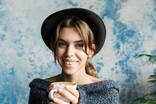 Feche o retrato de uma jovem vestida com um suéter e um chapéu na parede azul, segurando uma xícara de café quente