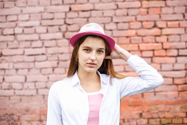 Feche o retrato de uma linda garota estilosa de chapéu perto da parede de tijolos rosa como pano de fundo