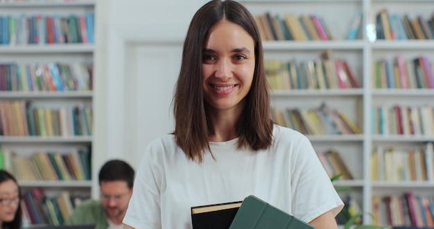 Feche o retrato de uma linda jovem segurando livro e tablet digital no fundo da estante da biblioteca olhando para a câmera Conceito de educação universitária