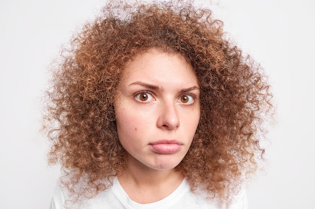 Foto feche o retrato de uma linda mulher europeia de cabelos cacheados olhando com grande admiração, não consigo entender algo tem uma pele limpa saudável cabelo cacheado espesso posa contra uma parede branca